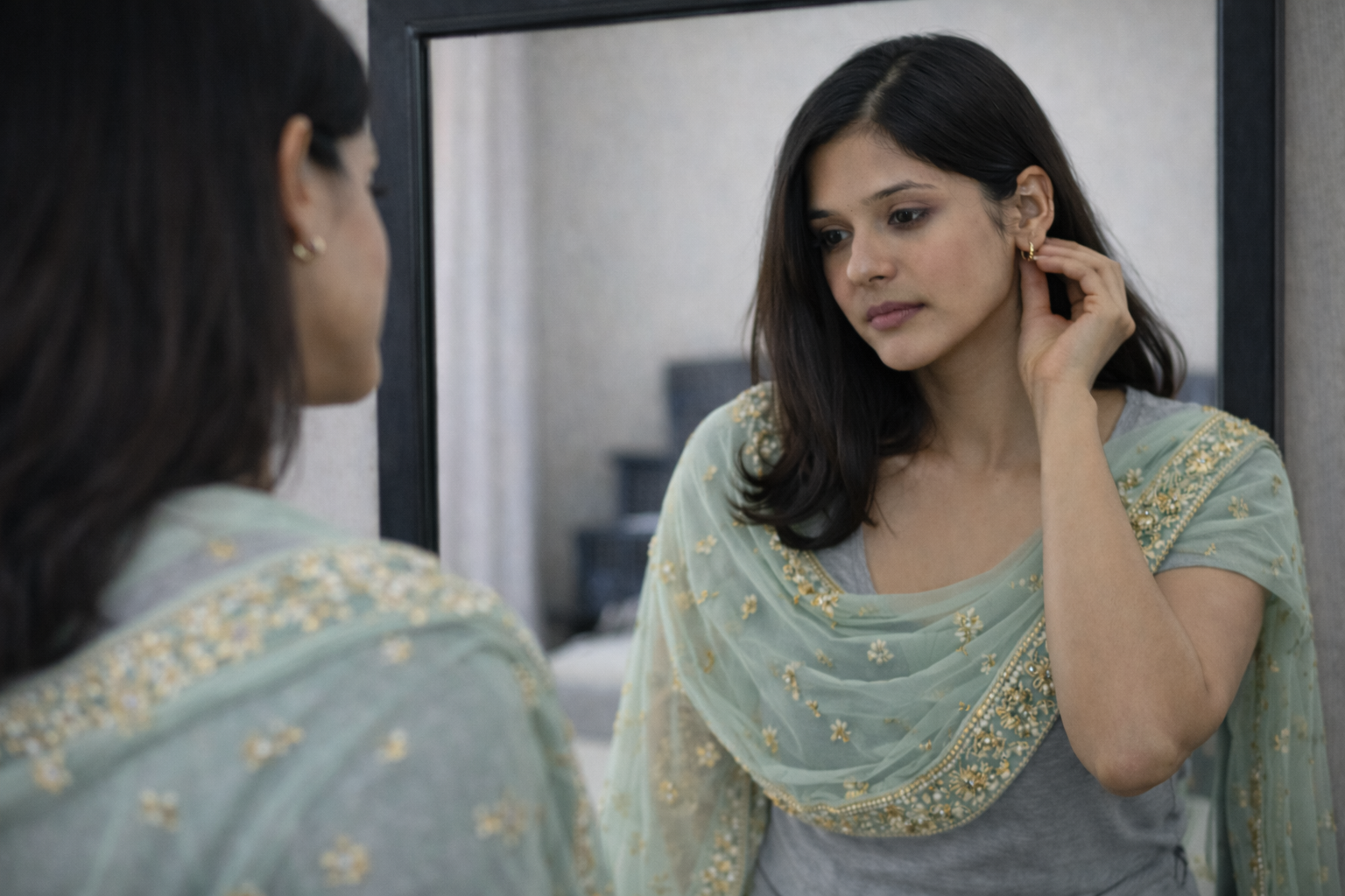 A young South Asian woman stands in front of a mirror, gently touching her ear while holding a mint-green, gold-embroidered dupatta over a grey top, thoughtfully considering which earrings to wear.